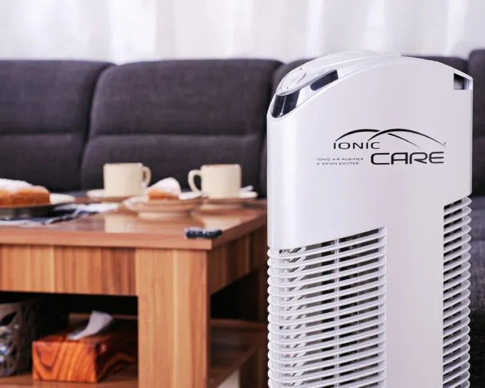 Čistička vzduchu Ionic-CARE v detailu u konferenčního stolku s kávou a koláčem, vedle tmavě šedé pohovky, barva perleťově bílá