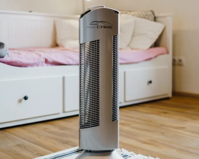 Čistička vzduchu Ionic-CARE ve světlém interiéru s postelí a modře pruhovanou předložkou, barva stříbrná
